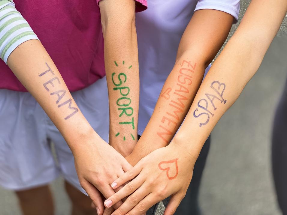 Vier Arme liegen übereinander, mit farbiger Schrift: „TEAM“, „SPORT“, „ZUSAMMEN“, „SPASS“ auf der Haut.