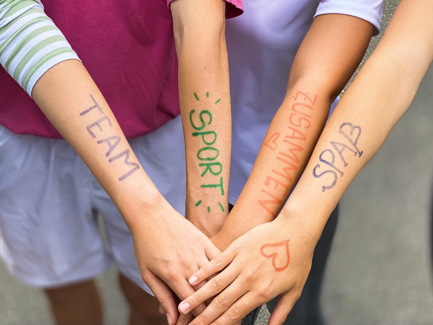 Vier Arme liegen übereinander, mit farbiger Schrift: „TEAM“, „SPORT“, „ZUSAMMEN“, „SPASS“ auf der Haut.